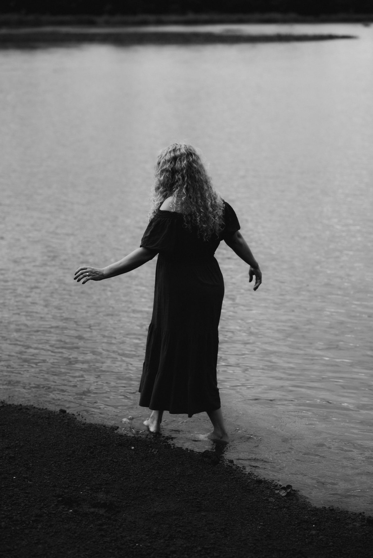 A woman in a long dress steps barefoot into the water, arms loose at her sides, seen from behind