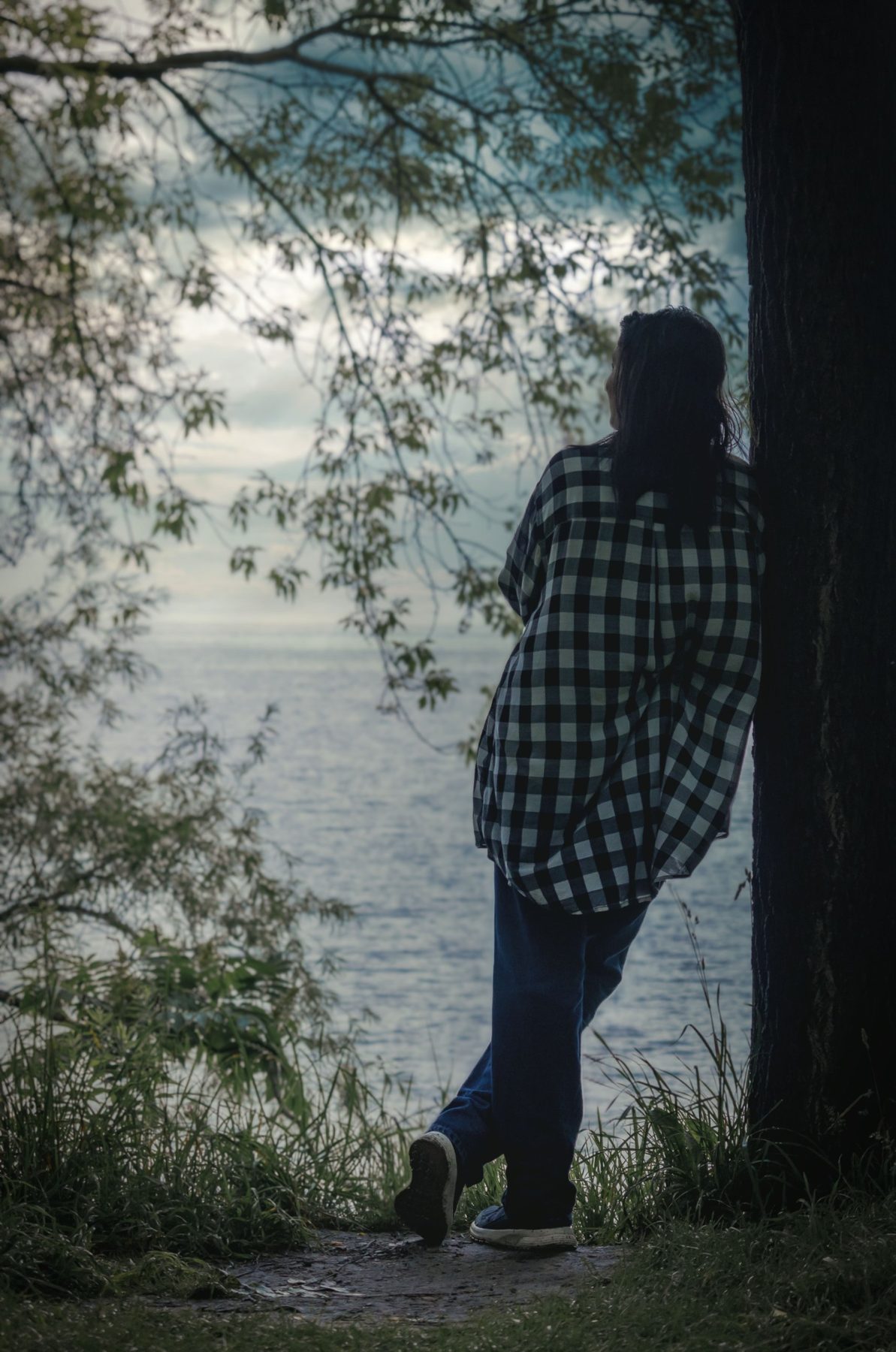 A person leaning against a tree, looking out over a quiet lake