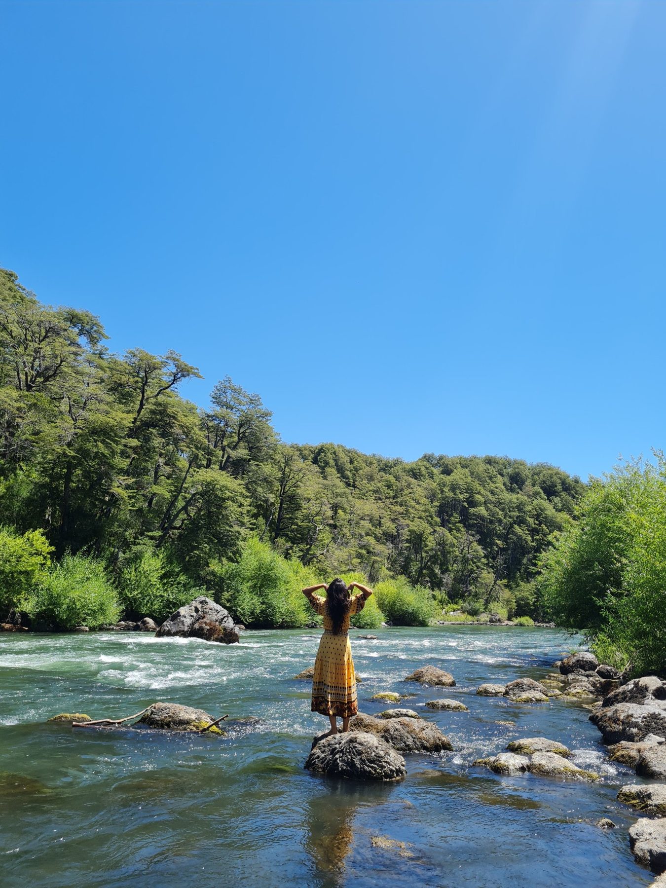 A woman standing on a rock in a sunlit river, arms raised, surrounded by green trees